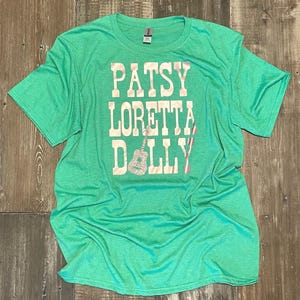 May include: A green t-shirt with the names PATSY, LORETTA, and DOLLY printed vertically in a white, bold font. A glittery guitar graphic is between LORETTA and DOLLY. The shirt is made of a soft material.