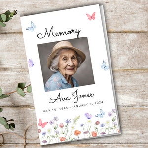 Op de afbeelding: Een wit gedenkboekje met een foto van een oudere vrouw met een hoed. Het boekje heeft de tekst "Memory" bovenaan en "Ava Jones" onder de foto. De data "May 15, 1945 - January 5, 2024" staan onder de naam gedrukt. Het boekje heeft een bloemmotief met vlinders aan de onderkant.