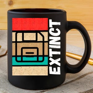 Op de afbeelding: Zwarte koffiemok met een retro-ontwerp met een floppy disk en de tekst "EXTINCT".