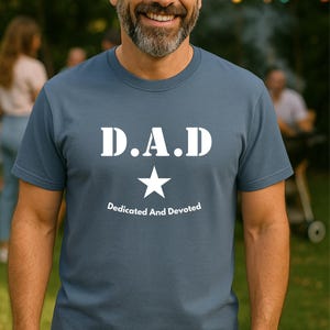 May include: A slate blue t-shirt with the letters "D.A.D" in bold white font, a white star, and the words "Dedicated And Devoted" below. The shirt is short-sleeved.