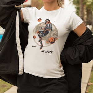 May include: White t-shirt with a graphic of an astronaut sitting on a moon with the text "Living My Space".