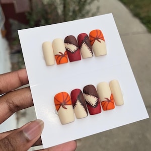 May include: Set of two press-on nail sets. Each set features five matte nails with fall-themed designs. Designs include pumpkins, stitched patterns, and solid colors in beige, orange, and burgundy.