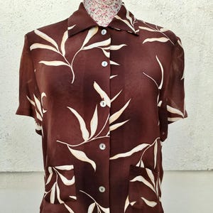 May include: A brown short-sleeved blouse with a white leaf pattern. The blouse has a collared neckline, button-down front, and two front pockets. The blouse is displayed on a mannequin.