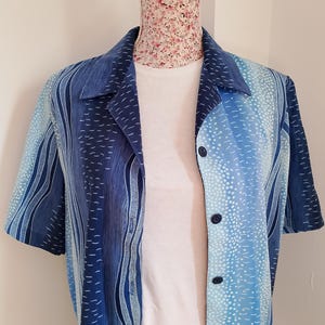 Puede incluir: Una camisa de manga corta con botones y un estampado azul y blanco. La camisa presenta un diseño degradado con rayas verticales y detalles de puntos. El cuello y los botones son de un tono azul más oscuro. La camisa se exhibe en un maniquí.