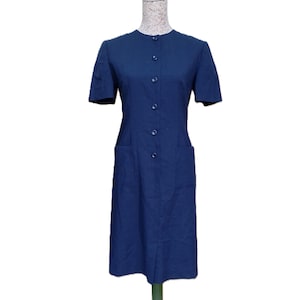 May include: A solid navy blue knee-length dress featuring short sleeves, a round neckline, and a front button-down closure with six matching buttons. The garment includes two large patch pockets on the front and a straight hemline with a center slit.