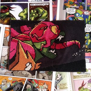 May include: A rectangular cross-stitch embroidery piece featuring a stylized, vibrant pink and green creature with a large yellow eye against a solid black background. The design is composed of visible, individual stitched squares.