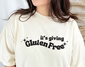 Op de afbeelding: Een wit T-shirt met de tekst "it's giving Gluten Free" in zwart met twee kleine sterren aan weerszijden van de tekst.