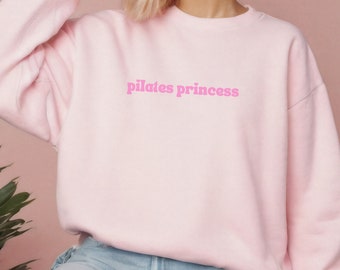 May include: A light pink sweatshirt with the text "pilates princess" in a bubblegum pink font. The long-sleeved crew neck top is made of a soft material. The sweatshirt is a casual style.