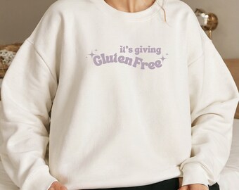 May include: White sweatshirt with a purple graphic that says "it's giving Gluten Free" with sparkles on either side of the text.