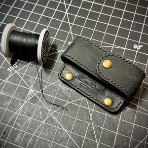 May include: A black leather needle case with two brass-colored snap closures and a spool of black thread. The needle case has the text "Monte Heather Handmade Leather Goods" embossed on it. The spool of thread is white and has black thread wrapped around it.