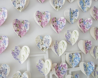 Può includere: Decorazioni a forma di cuore di carta con disegni floreali nei toni del viola, blu e bianco. I cuori sono sospesi su uno sfondo bianco, creando un'estetica delicata e romantica. I cuori sono fatti di pagine di libri.