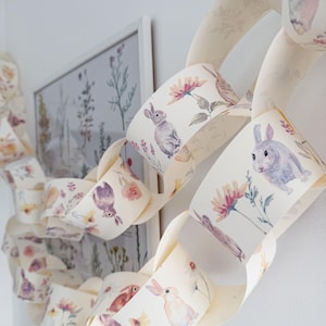 May include: A paper chain garland with watercolor illustrations of bunnies, flowers, and foliage. The links are cream-colored with pastel accents of pink, purple, and yellow. A framed botanical print is in the background.