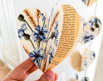 Puede incluir: Guirnalda de papel hecha a mano con forma de corazón. Cada corazón presenta un diseño floral con flores azules y espigas de trigo, sobre un fondo de páginas de libro envejecidas. La guirnalda está suspendida, creando un elemento decorativo delicado.