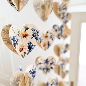 May include: Decorative heart-shaped garlands. Each heart is crafted from pages of a book and adorned with floral patterns in white, blue, and orange. The hearts are suspended, creating a whimsical and romantic display.