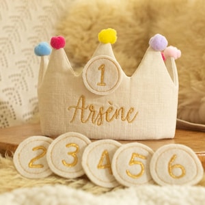 Peut inclure: Une couronne en lin blanc avec un numéro un brodé en or et le nom "Arsène" dessus. La couronne est décorée de pompons colorés et est entourée de six cercles en feutre blanc avec des numéros deux à six brodés en or.