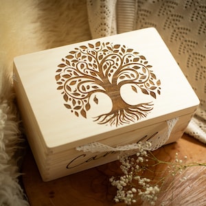 Peut inclure: Une boîte en bois avec un motif d'arbre de vie gravé sur le couvercle. La boîte est de couleur brun clair et possède une bordure en dentelle blanche. La boîte est posée sur une surface en bois.