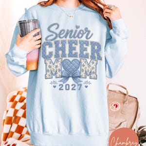 以下が含まれることがあります： キルティングと花柄の「Senior CHEER MOM」という文字がプリントされたライトブルーのクルーネックトレーナー。ハート型の「O」の下には青いリボンがあり、その下に2027という年号が記されています。デザインには小さなハートと葉のアクセントが含まれています。