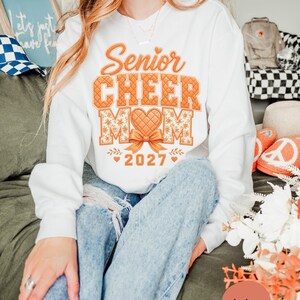 以下が含まれることがあります： Senior CHEER MOM 2027と書かれたオレンジ色のグラフィックプリントが施された白い長袖スウェットシャツ。デザインには、キルティングされたハート、リボン、オレンジ色の花柄が含まれており、衣服の胸の中央に配置されています。