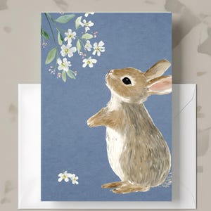 May include: A greeting card featuring a watercolor illustration of a brown rabbit looking up at white flowers on a blue background. The card is paired with a white envelope, perfect for sending a special message.