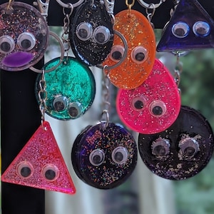 May include: A collection of colorful, glittery resin keychains in various shapes, including circles, ovals, and triangles. Each keychain features googly eyes for a whimsical touch and is attached to a silver keyring.