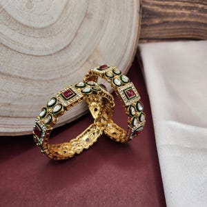 May include: Two gold bangles with intricate designs. The bangles are adorned with red and white stones, and green accents.