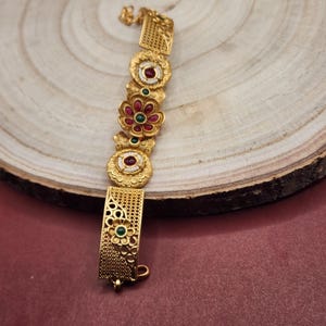 Könnte beinhalten: Goldfarbenes Armband mit floralen und geometrischen Mustern. Das Armband hat kreisförmige und rechteckige Glieder, die mit roten und grünen Edelsteinen verziert sind. Die floralen Elemente haben rote Steine in der Mitte, umgeben von weißem Emaille.