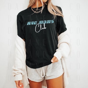 Op de afbeelding: Zwart T-shirt met de tekst "BAD MOMS Club" in blauw en wit. Het shirt wordt gedragen met grijze shorts en een crèmekleurig vest. Het shirt heeft een casual stijl.