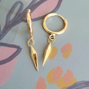 May include: A pair of gold hoop earrings featuring dangling, pointed, spike-shaped charms. Each earring consists of a circular hoop with a hinged closure and a small jump ring connecting to a faceted, metallic, cone-like pendant that hangs vertically.