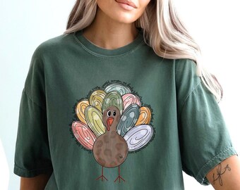 May include: Moss green t-shirt featuring a colorful turkey graphic. The turkey has a brown body, a red wattle, and a fan of pastel-colored feathers. The shirt is a relaxed fit and has short sleeves. The word "MOSS" is in a green circle.