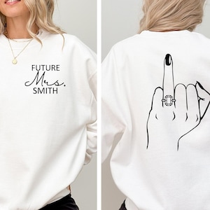 May include: White sweatshirt with the text "FUTURE Mrs. SMITH" on the front and a black and white illustration of a hand giving the middle finger with a ring on the ring finger on the back.