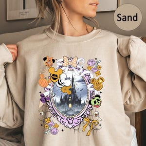 May include: Sand-colored sweatshirt featuring a Halloween-themed graphic. The design includes a castle, pumpkins, Mickey Mouse-inspired characters, and floral elements within an ornate oval frame. The overall aesthetic is whimsical and festive.