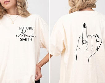 May include: Ivory oversized t-shirt with the text "FUTURE Mrs. SMITH" on the front and a black line drawing of a hand giving the middle finger with a ring on the finger on the back.