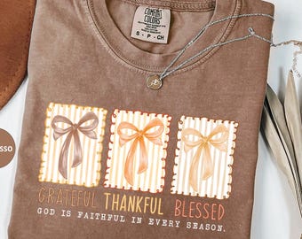 May include: Espresso-colored t-shirt with a graphic of three bows in frames, with the words "Grateful, Thankful, Blessed." The text "God is faithful in every season" is also printed on the shirt. A silver necklace and a brown hat are also visible.