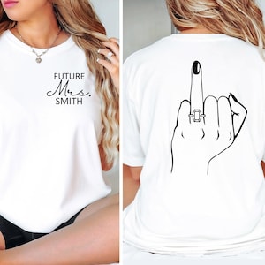 May include: White t-shirt with the text "FUTURE Mrs. SMITH" on the front and a black line drawing of a hand with a ring on the middle finger giving the middle finger on the back.
