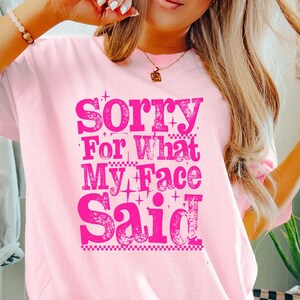 May include: A light pink t-shirt with the text "Sorry For What My Face Said" in a bright pink, stylized font. The shirt is a casual style, suitable for everyday wear. The word "Blossom" is in a pink circle at the bottom right.