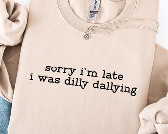 May include: A beige sweatshirt with the text "sorry i'm late i was dilly dallying" in black. The sweatshirt is folded with a pair of blue jeans with distressed details. A casual outfit.