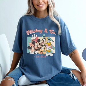 May include: A blue t-shirt with a vintage-style graphic of Mickey Mouse and friends. The graphic features Mickey Mouse, Minnie Mouse, Donald Duck, Goofy, and Pluto. The text on the shirt reads "Mickey & Co. Est. 1928".