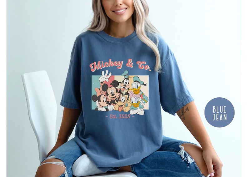 May include: A blue t-shirt with a vintage-style graphic of Mickey Mouse and friends. The graphic features Mickey Mouse, Minnie Mouse, Donald Duck, Goofy, and Pluto. The text on the shirt reads "Mickey & Co. Est. 1928".