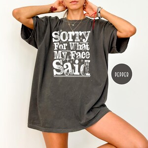 May include: Dark gray oversized t-shirt with the text "Sorry For What My Face Said" in white. The shirt has a vintage, worn look and a round "Pepper" label.