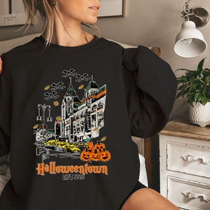 Puede incluir: Sudadera negra con un estampado gr&aacute;fico de una ciudad fantasmal con el texto "Halloweentown EST. 1998" y una pila de calabazas naranjas.