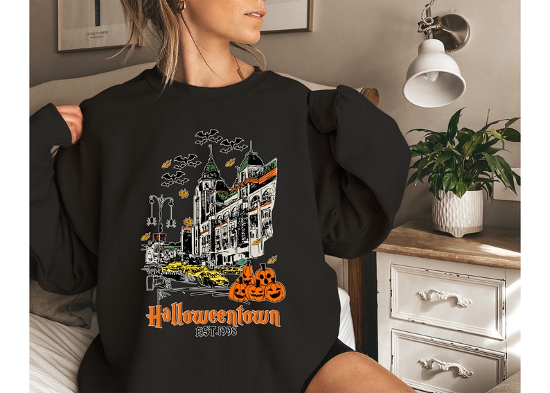 Puede incluir: Sudadera negra con un estampado gr&aacute;fico de una ciudad fantasmal con el texto "Halloweentown EST. 1998" y una pila de calabazas naranjas.