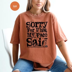 May include: A rust-colored t-shirt with the text "Sorry For What My Face Said" in a bold, black, decorative font. The shirt has short sleeves and a relaxed fit. The model is wearing blue jeans.