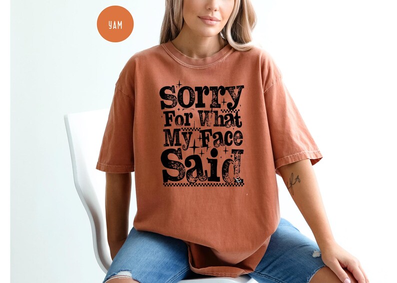 May include: A rust-colored t-shirt with the text "Sorry For What My Face Said" in a bold, black, decorative font. The shirt has short sleeves and a relaxed fit. The model is wearing blue jeans.