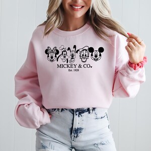 May include: A pale pink sweatshirt with black and white cartoon character faces, including Minnie Mouse, Goofy, Pluto, Donald Duck, and Mickey Mouse. The text "Mickey & Co. Est. 1928" is printed below the characters.