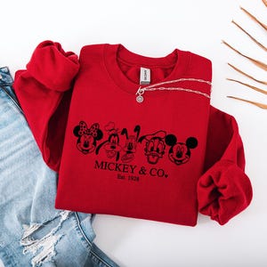 May include: A red sweatshirt with a black graphic of Mickey Mouse, Minnie Mouse, Goofy, Pluto, and Donald Duck. The text "Mickey & Co. Est. 1928" is below the characters. A silver necklace and blue jeans are also in the image.