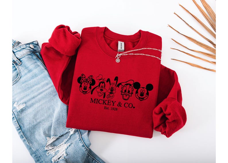May include: A red sweatshirt with a black graphic of Mickey Mouse, Minnie Mouse, Goofy, Pluto, and Donald Duck. The text "Mickey & Co. Est. 1928" is below the characters. A silver necklace and blue jeans are also in the image.