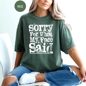 May include: A moss green t-shirt with the text "Sorry For What My Face Said" in a white, bold, and playful font. The shirt is worn with blue jeans. The overall style is casual and relaxed.