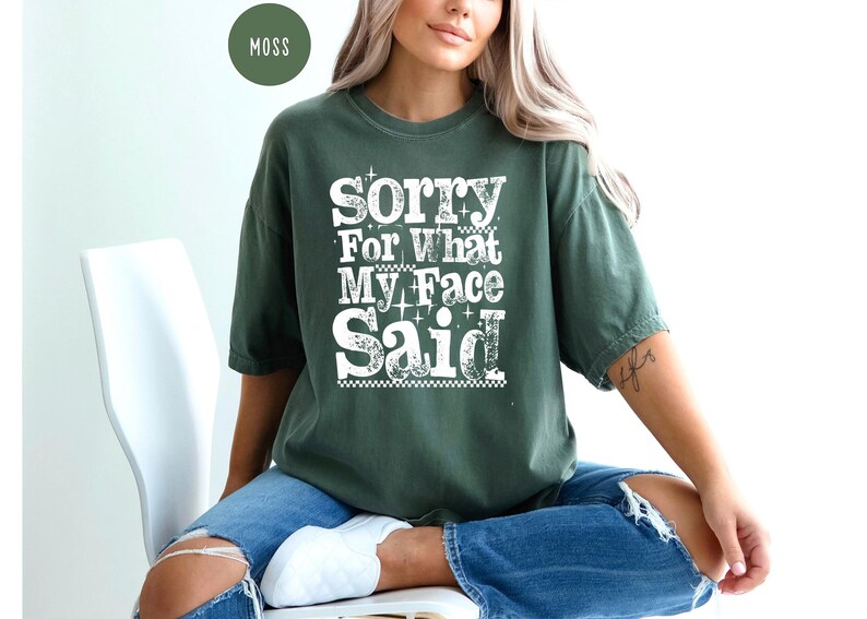 May include: A moss green t-shirt with the text "Sorry For What My Face Said" in a white, bold, and playful font. The shirt is worn with blue jeans. The overall style is casual and relaxed.