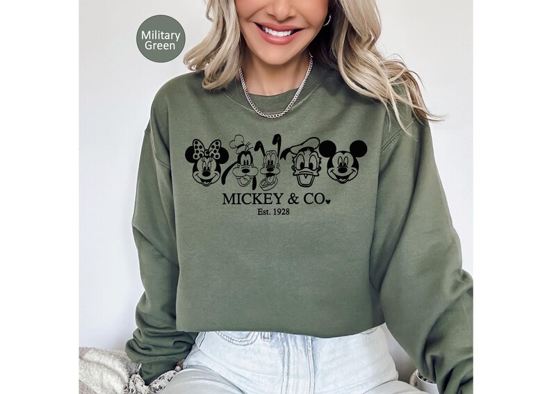 May include: A military green sweatshirt with a black graphic of Mickey Mouse, Minnie Mouse, Goofy, Pluto, Donald Duck, and Mickey Mouse. The text "Mickey & Co." and "Est. 1928" are printed below. A casual style sweatshirt.