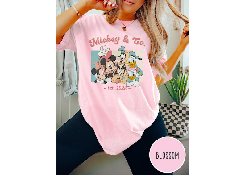 May include: Pink t-shirt with a vintage-style graphic of Mickey Mouse, Minnie Mouse, Donald Duck, and Goofy. The graphic includes the text "Mickey & Co. - Est. 1928 - "
